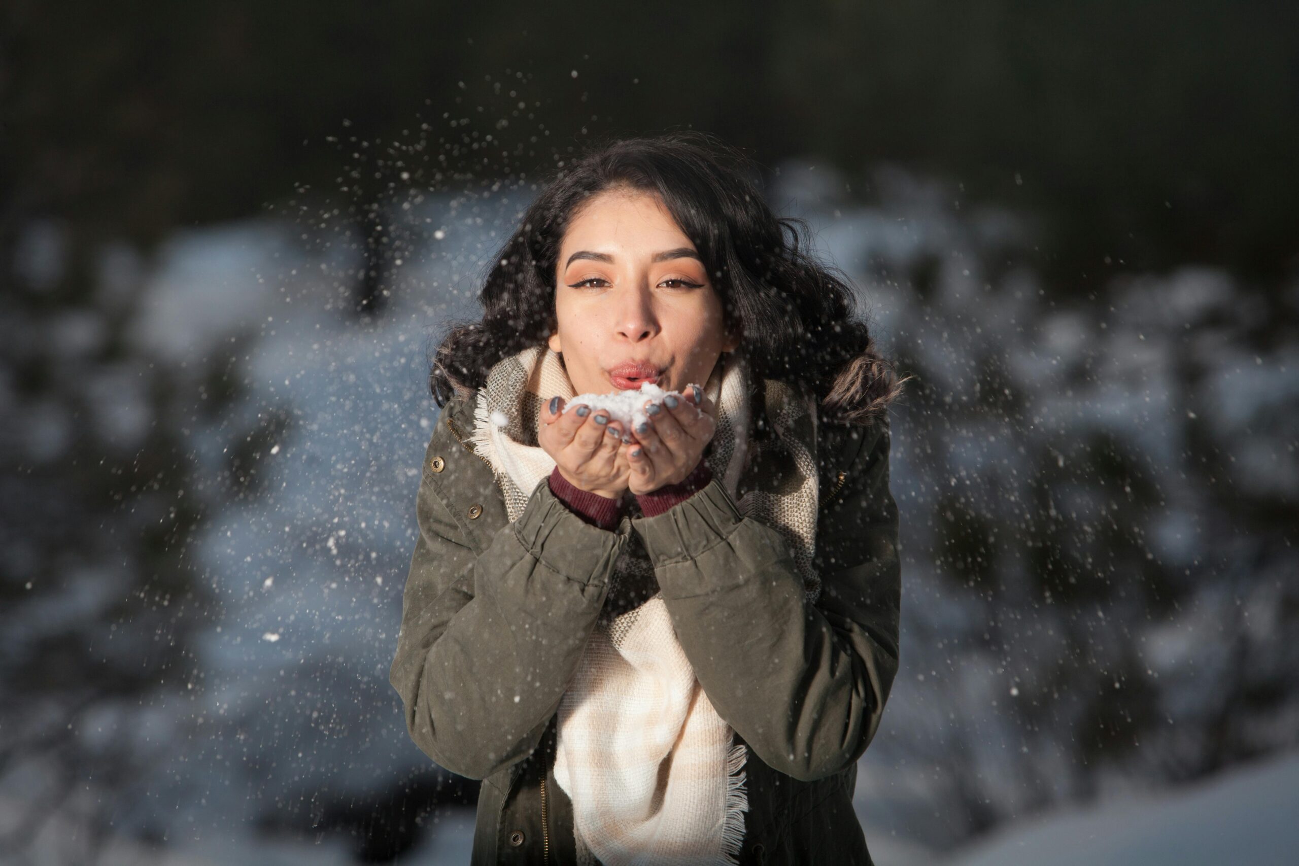 Women wearing a casual winter outfit. Ths woman is standing in the snow and is blowing a pile of snow to the photographer.