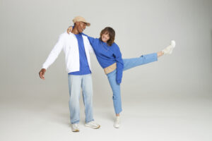 Male and female models wearing matching blue hoodies. Both are wearing denim jeans. Both are smiling and the girl is kicking her left leg in the air.
