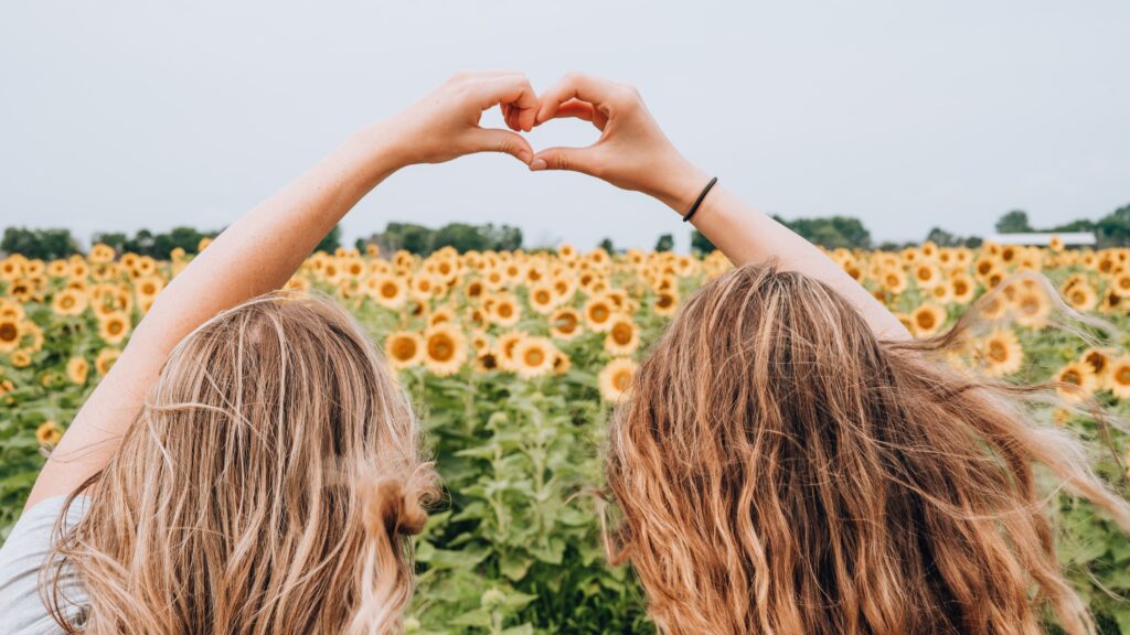 Two girls making the heart sign with their hands in the air above a field of sunflowers.