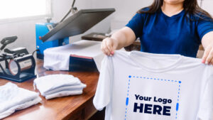 A woman standing by a DTF printer, holding up a t-shirt with "your logo here" written on it